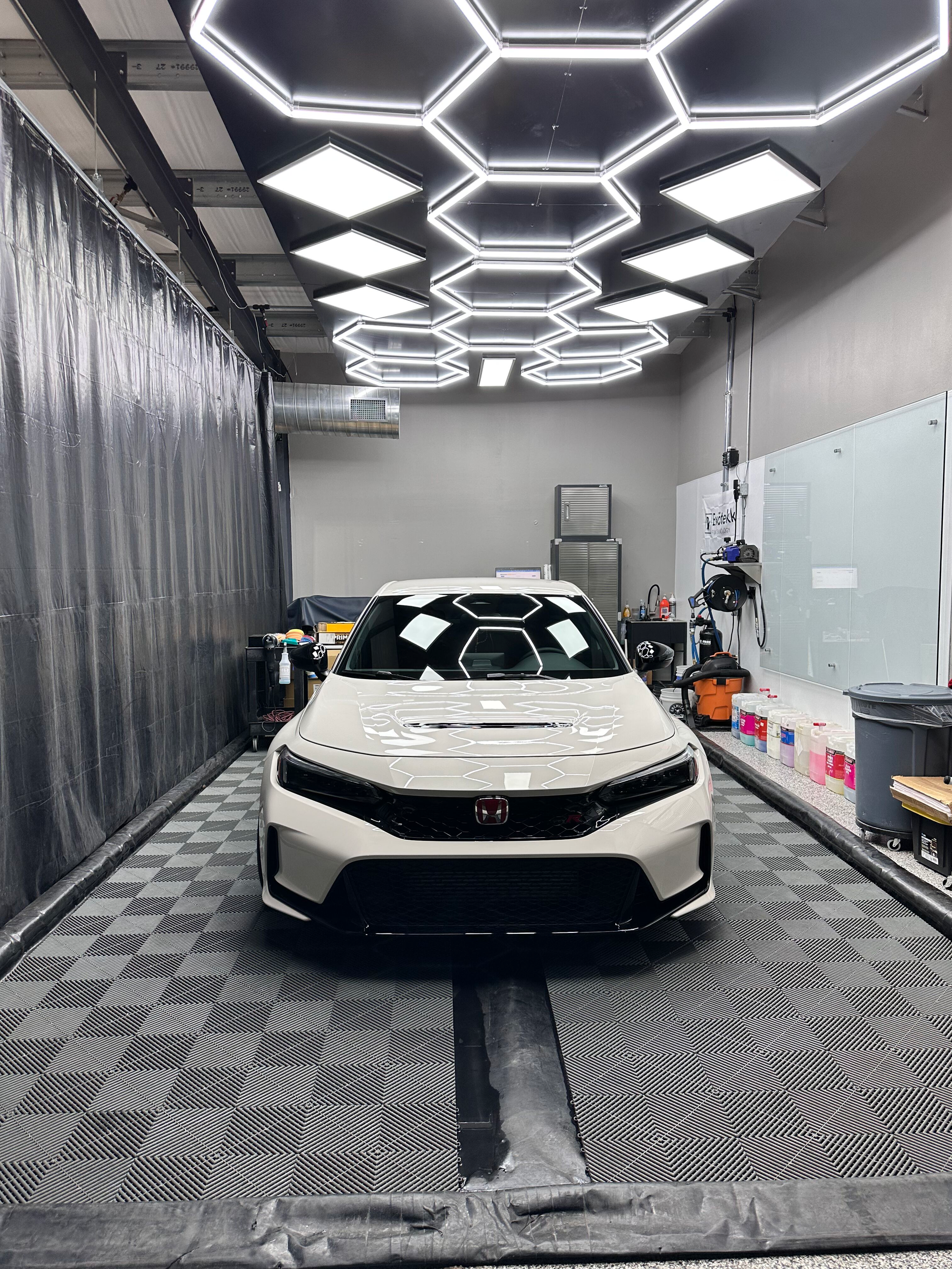 A white car parked in a detailing bay with hexagon ceiling lights. The floor has a patterned design, and the walls are gray.