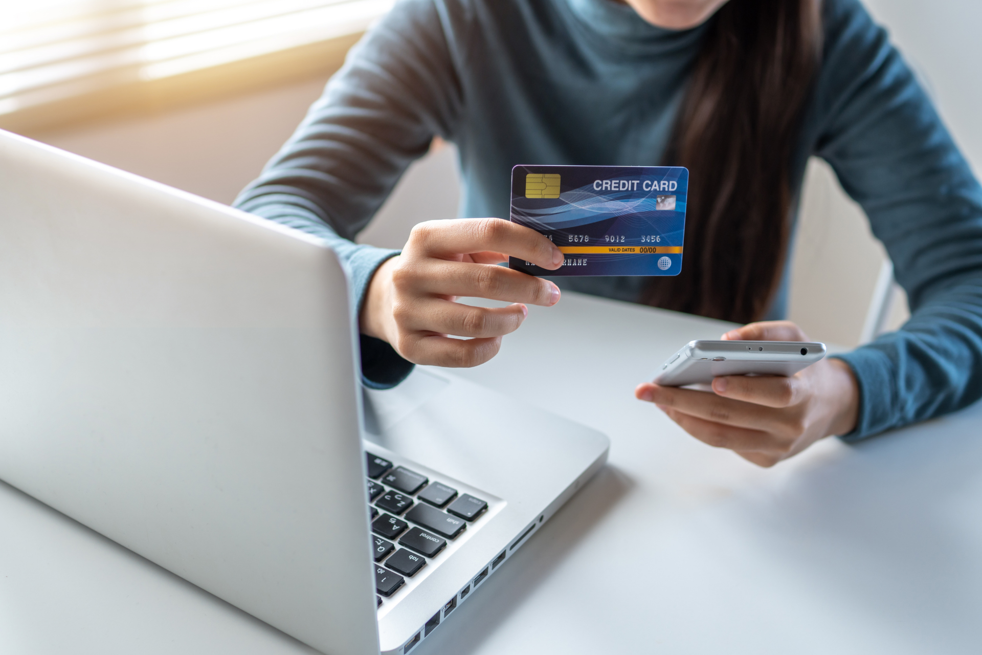 A person in a blue sweater holds a credit card and a smartphone while sitting in front of a laptop on a desk.