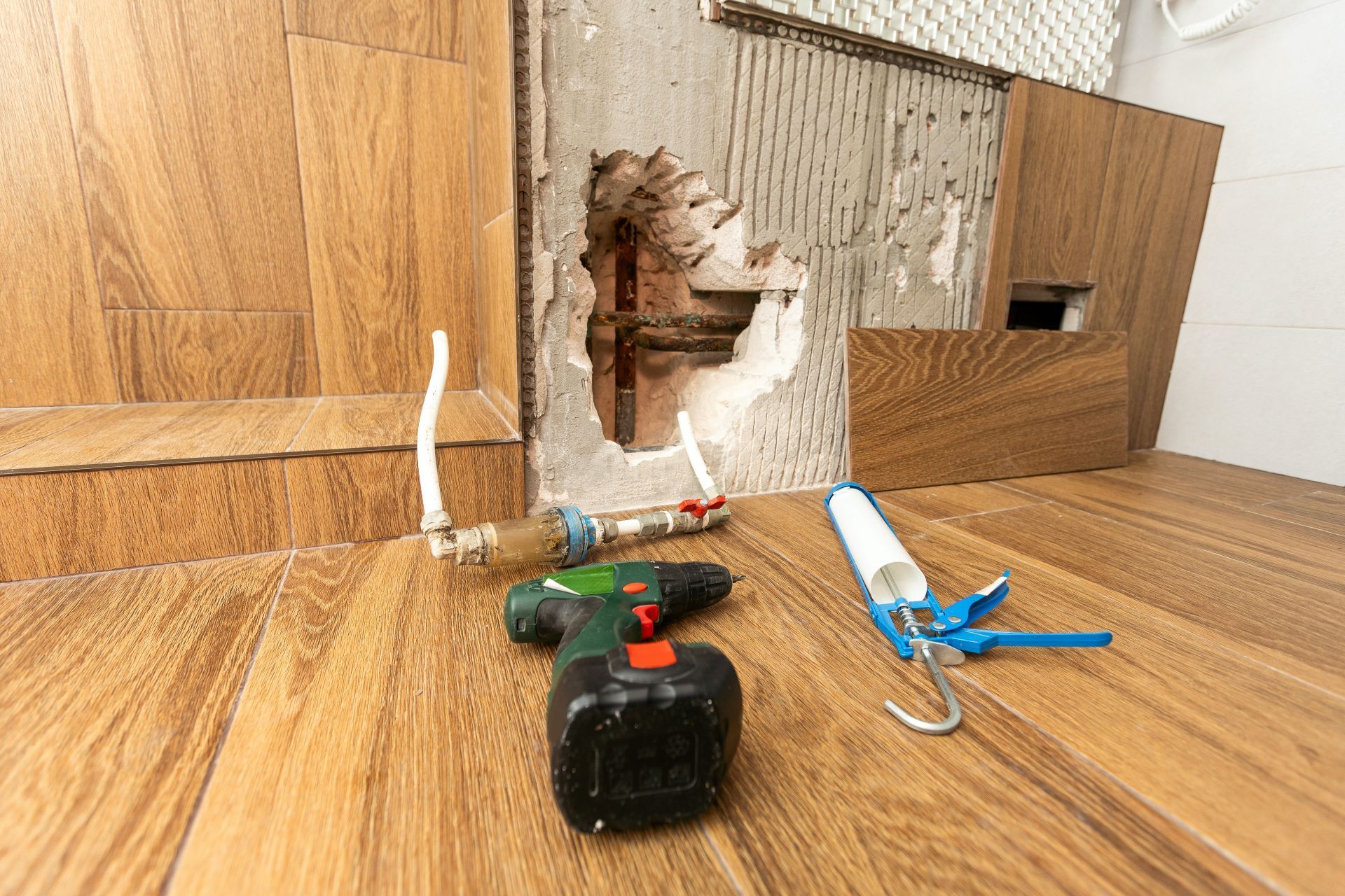 A construction scene with a hole in a tiled wall, a drill, and a caulk gun resting on a wood-look floor.
