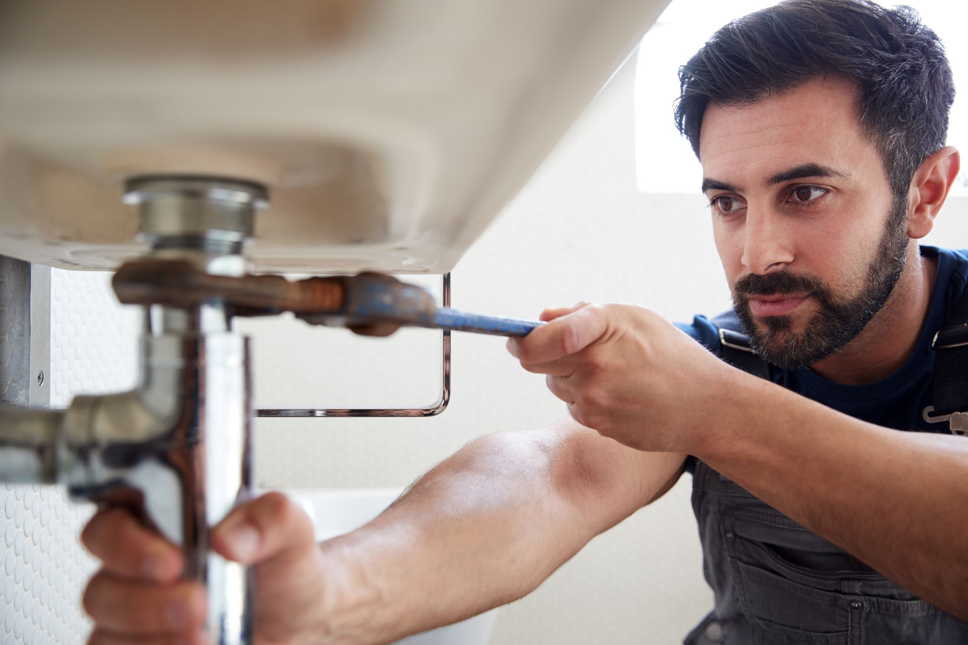 Plumber tightening pipe under a white sink with a wrench. The setting is indoors, near a wall.