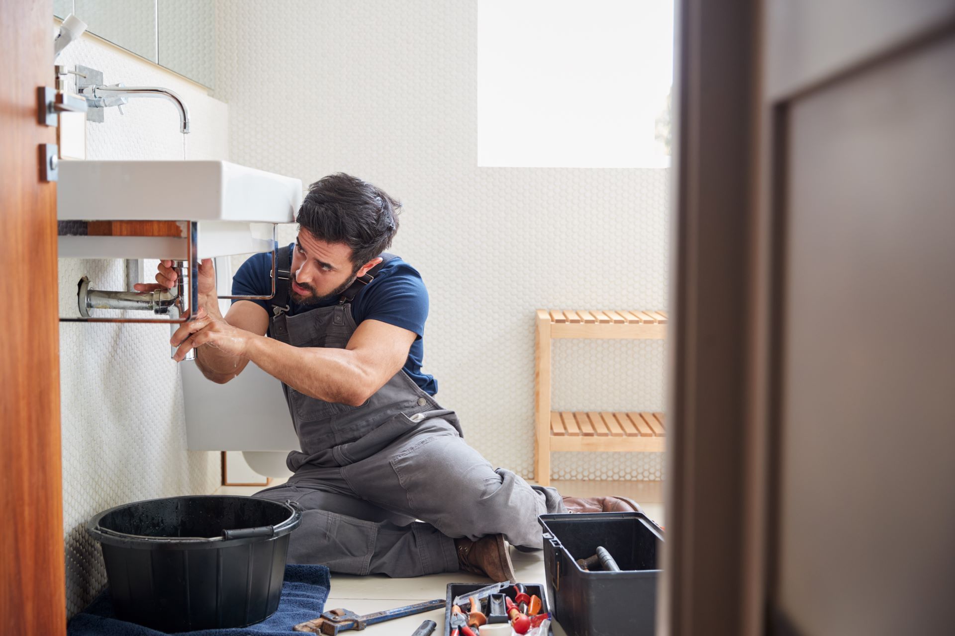 Technician repairing a sink pipe under a bathroom vanity, with tools and parts nearby.