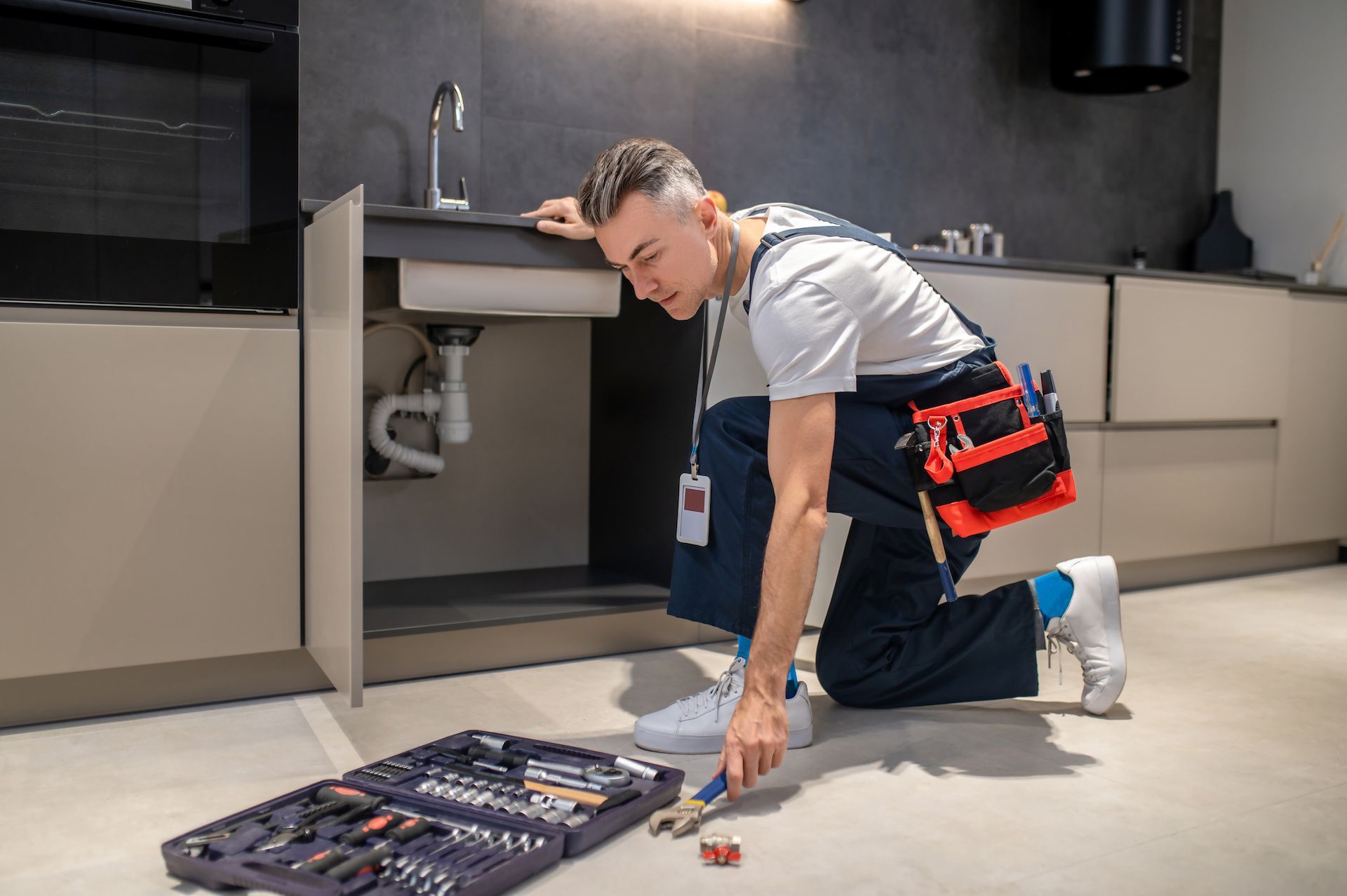 A man is kneeling on the floor fixing a sink in a kitchen.