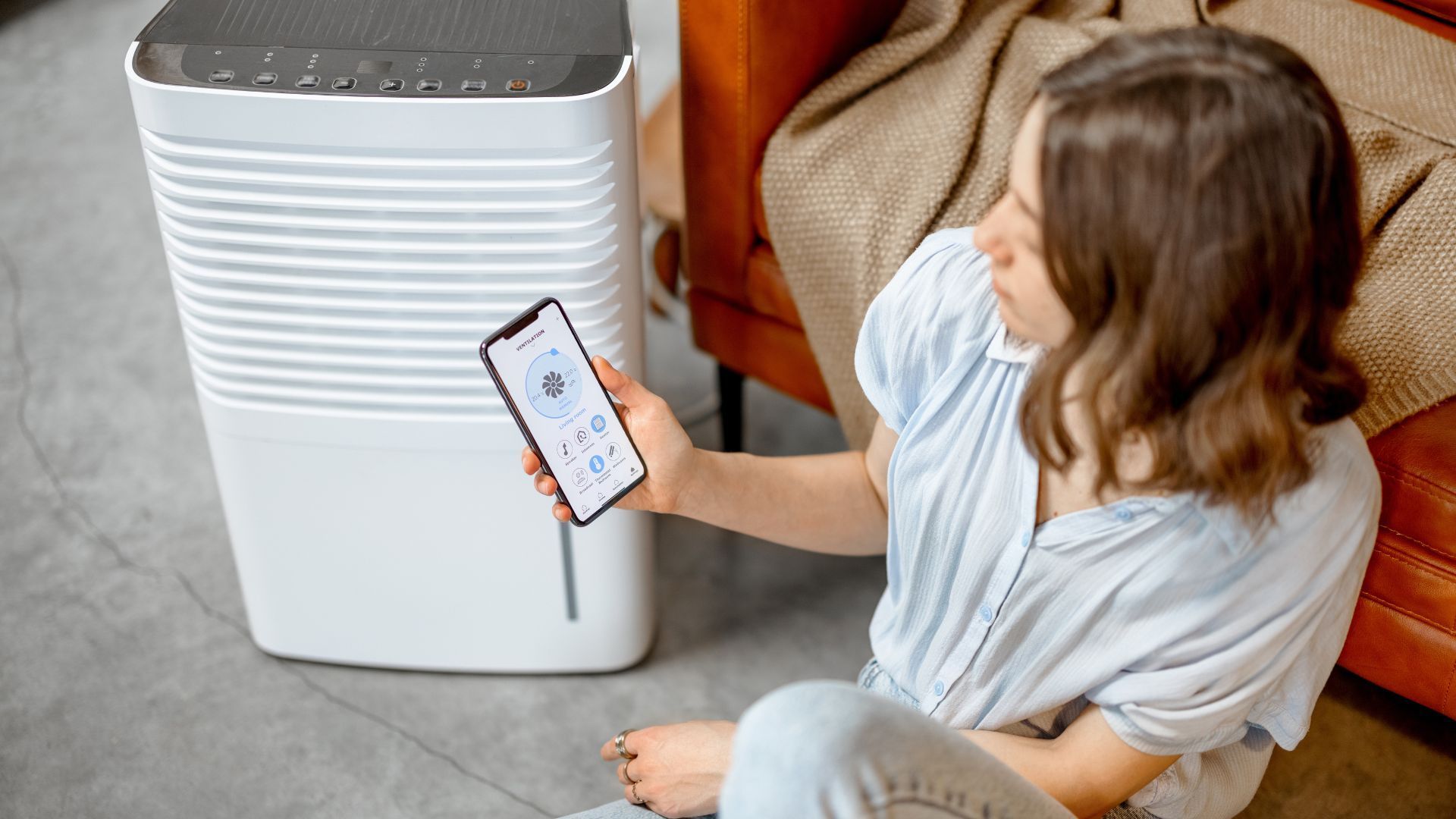 A woman is sitting on the floor holding a cell phone in front of a dehumidifier.