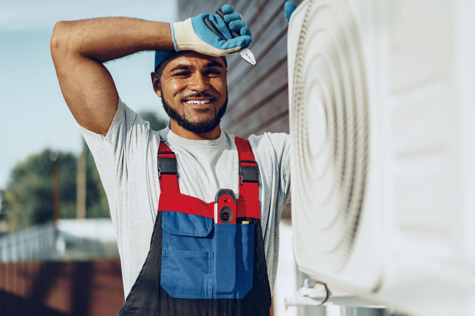HVAC technician wiping sweat, smiling, and leaning on an air conditioning unit outside.