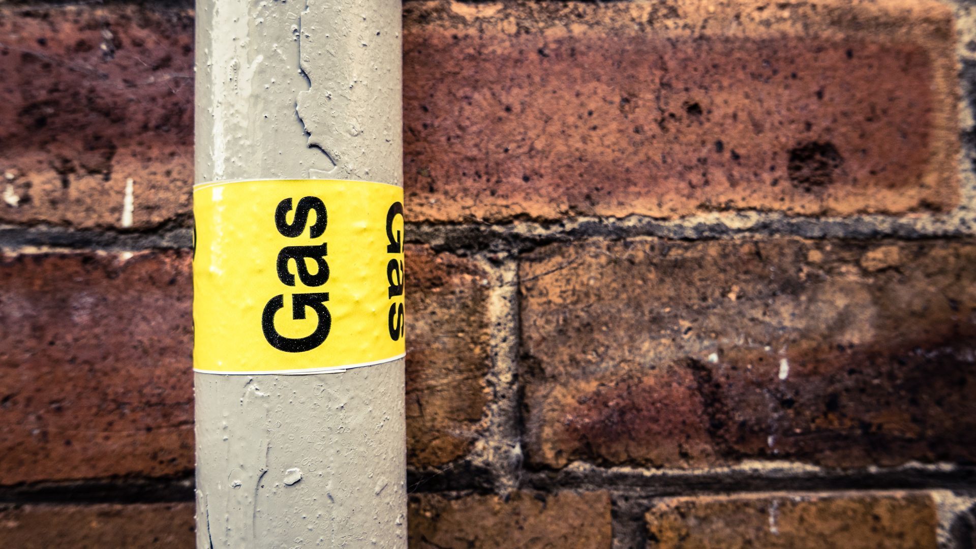 A gas pipe with a yellow sticker on it is against a brick wall.