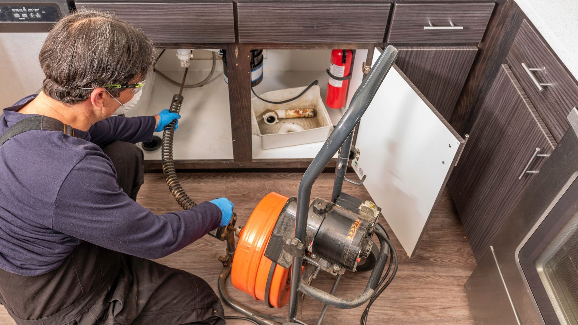 A man is using a machine to clean a drain under a sink in a kitchen.