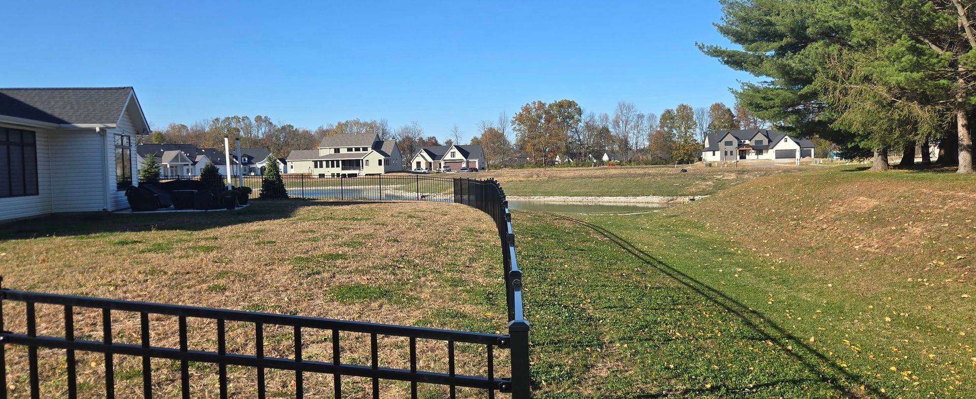 Grassy neighborhood field with a black fence in the foreground and houses under a clear blue sky