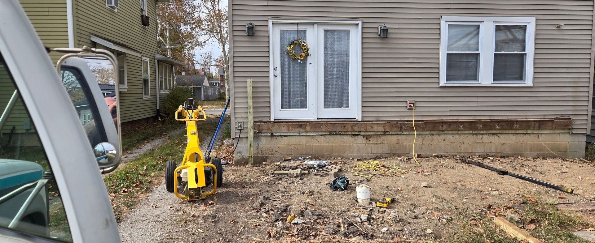 Yellow mini excavator in a dirt yard beside a beige house, clearing debris near the wall.