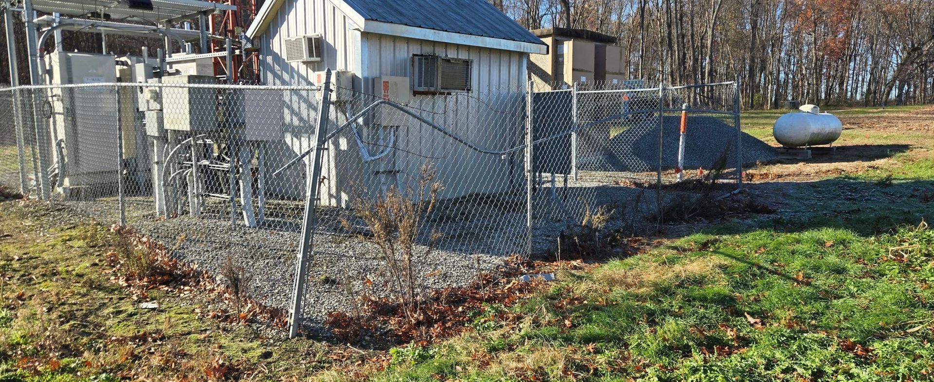 Chain-link fenced yard with a small white house, blue shed, and white propane tank in autumn sunlight