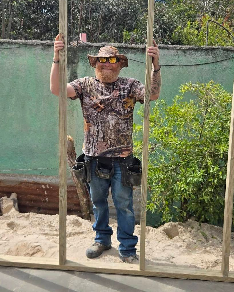 A person in a camouflage t-shirt, hat, and tool belt stands between two wooden vertical studs outdoors in the sand.
