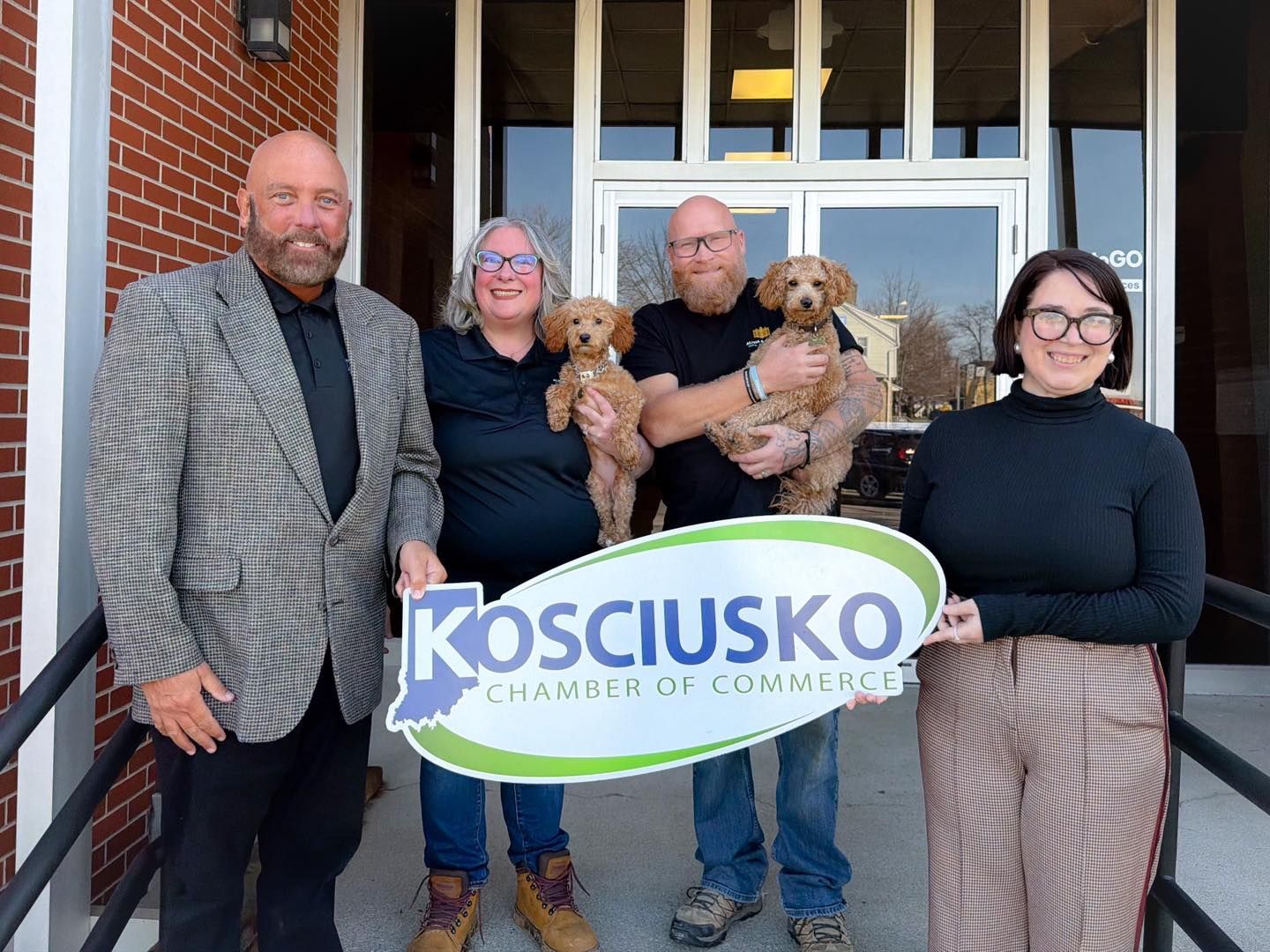Four people stand outside a brick building, two holding small dogs, while two hold a Kosciusko Chamber of Commerce sign.
