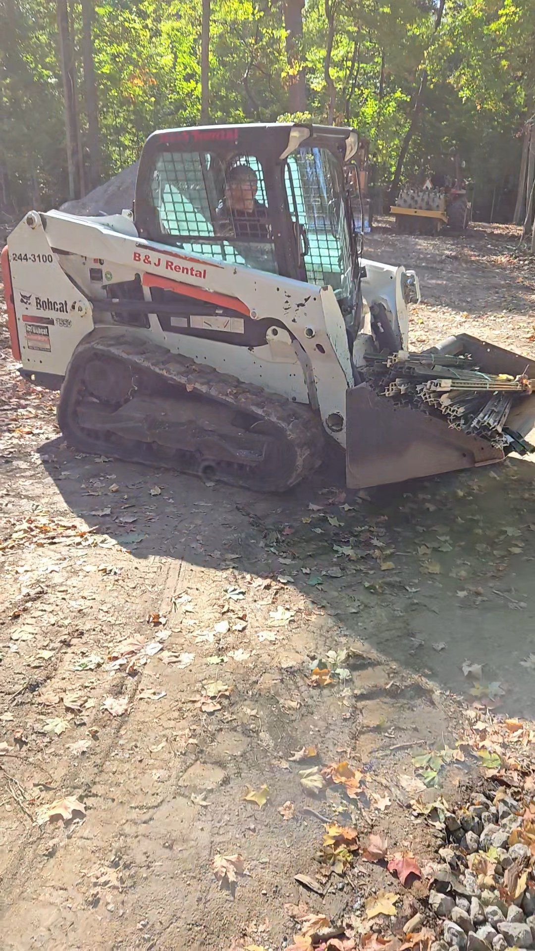 White skid-steer loader on a leaf-covered dirt path in a wooded area
