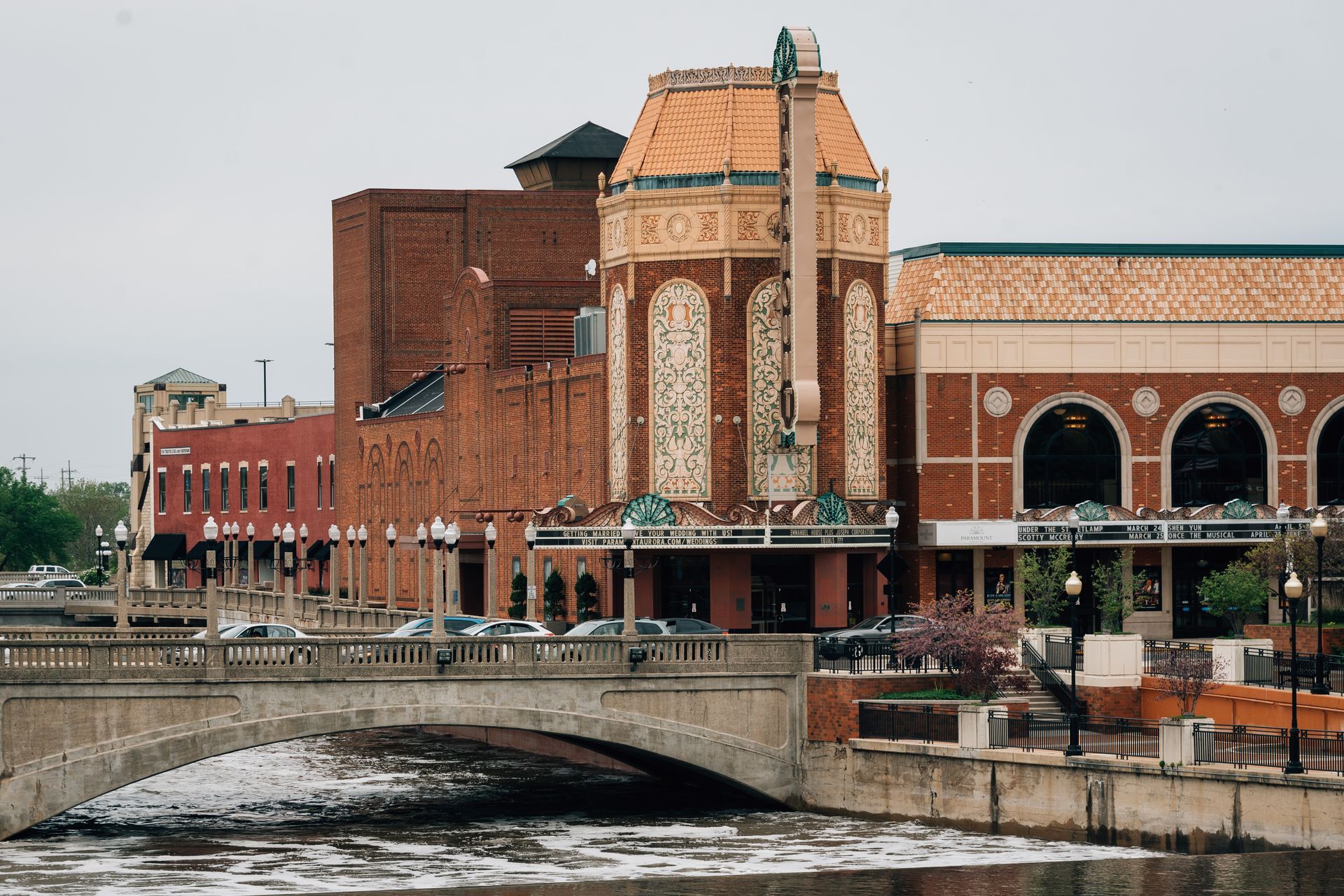 Brick buildings, including a theater, by a bridge over a river; overcast sky.
