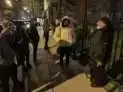 People stand on a city street at night, lit by streetlights. One person wears a yellow hooded jacket.