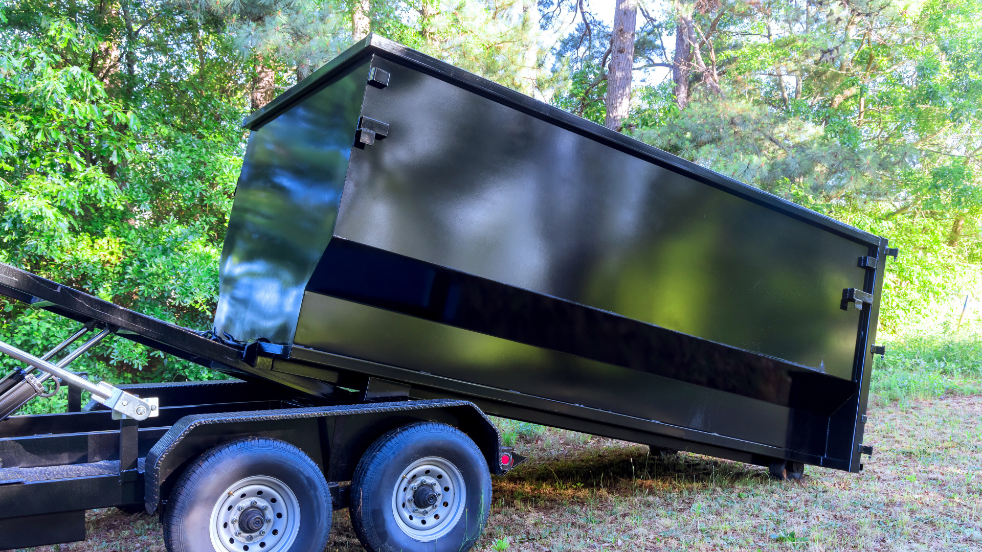 Black dumpster on a trailer, tilted upwards, outdoors in front of trees.