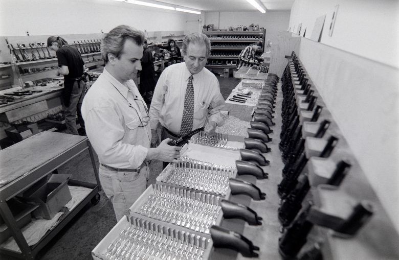 Two men inspecting handguns in a factory setting. Many assembled guns are visible on a rack.