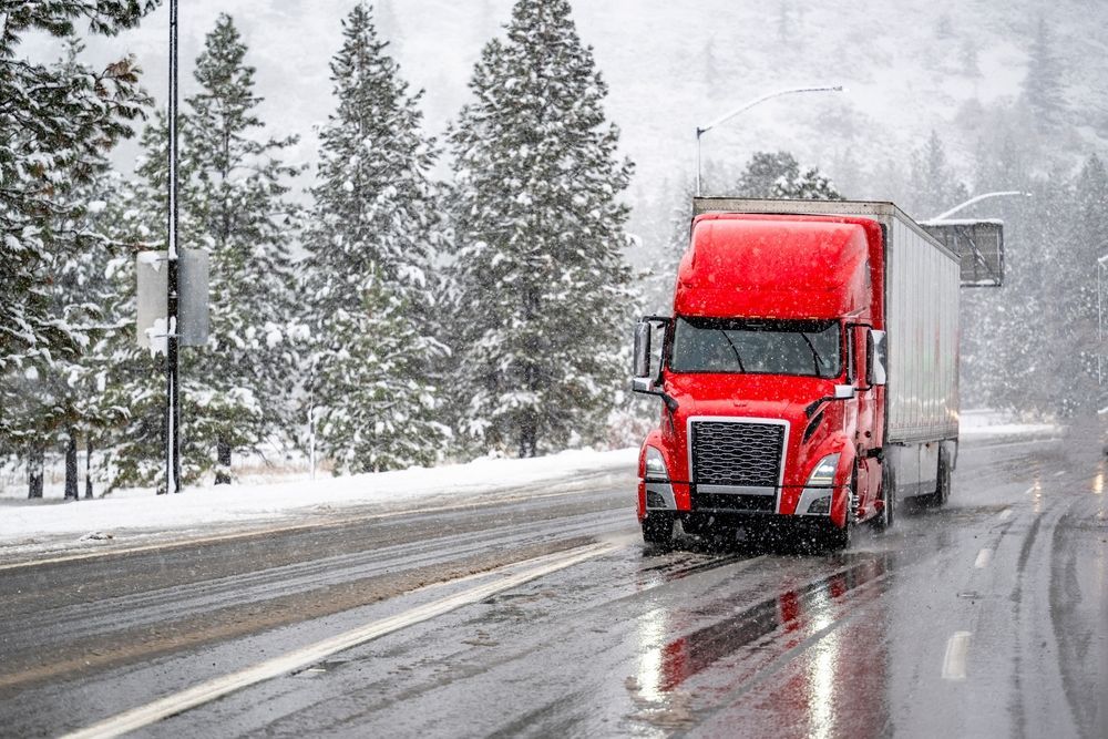 Red semi-truck driving on a snowy road with pine trees and a misty winter backdrop