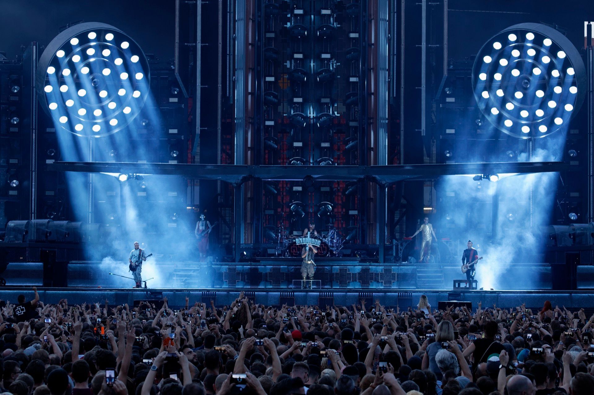 Band sul palco, immersa in una luce blu, davanti a una grande folla con le mani alzate.