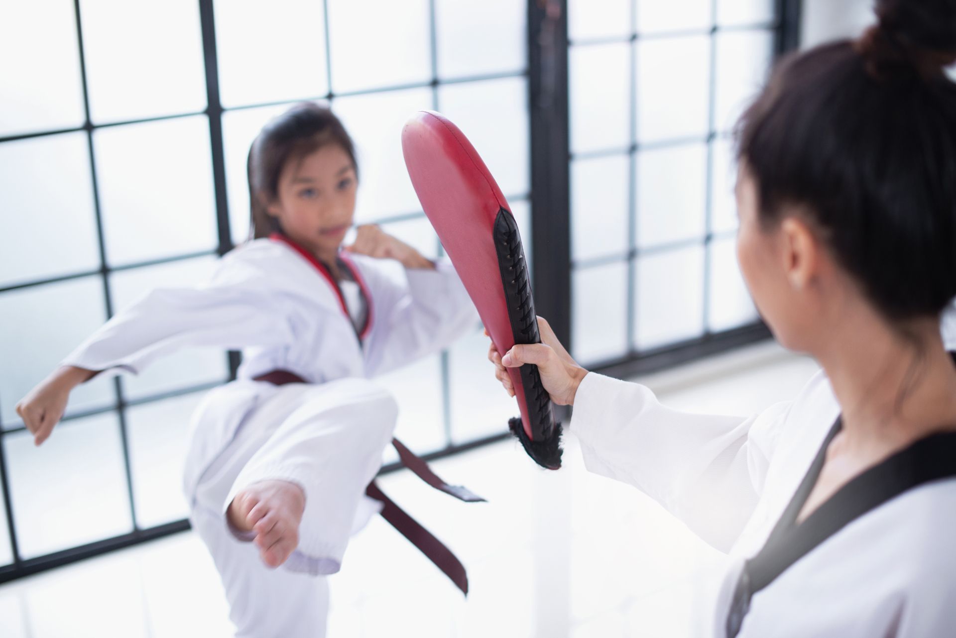 Girl in a white uniform kicking a red and black target held by a person in a white uniform.