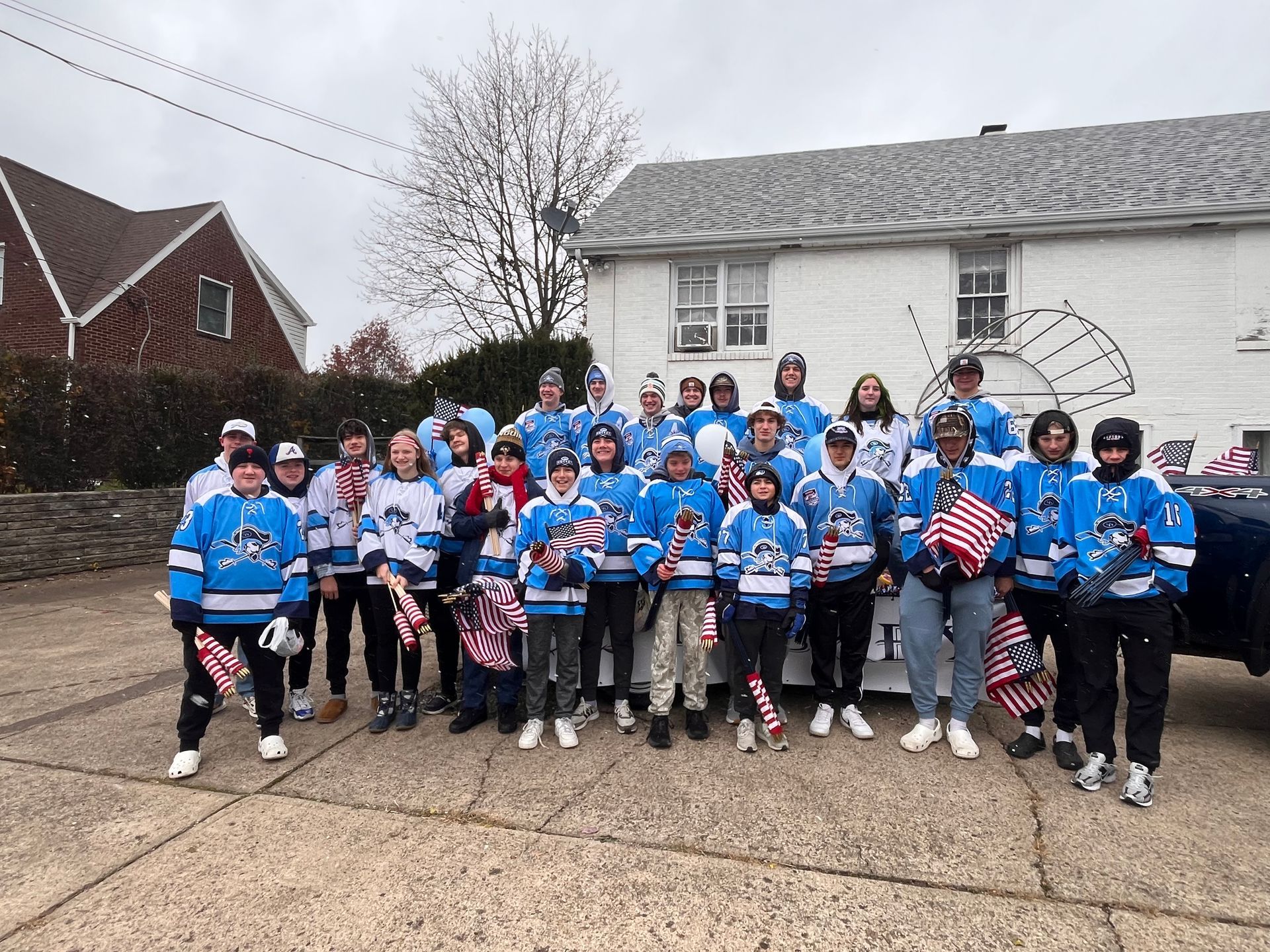 A large group in matching light blue and white sports jerseys pose together outside a white building, holding flags.