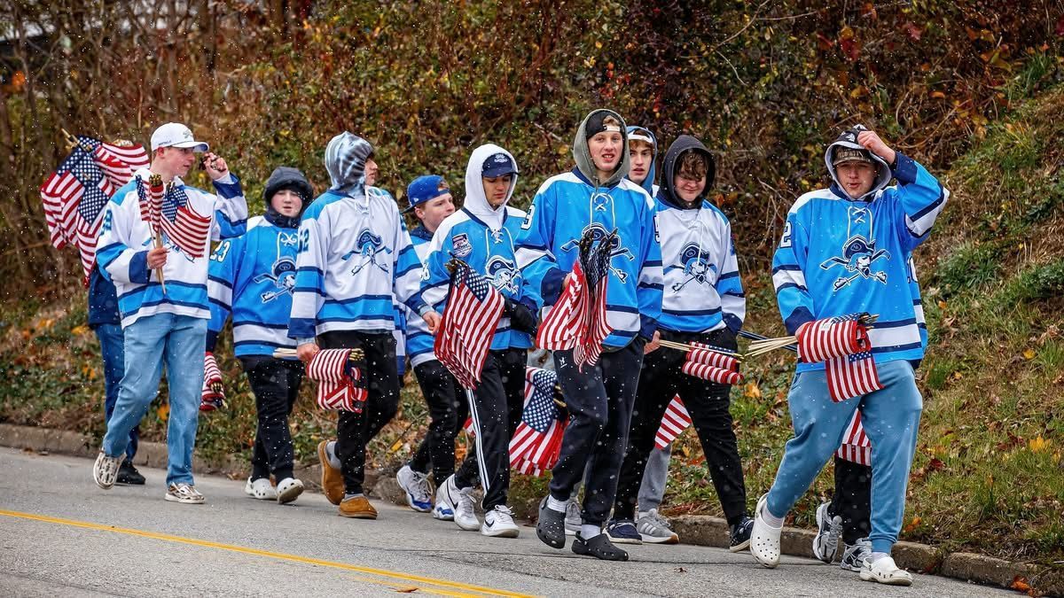 A group of people in matching light blue sports jerseys walk along a road, carrying small American flags.