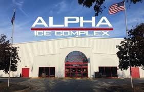 Exterior view of Alpha Ice Complex with a red entrance, white siding, and American flag on a sunny day.