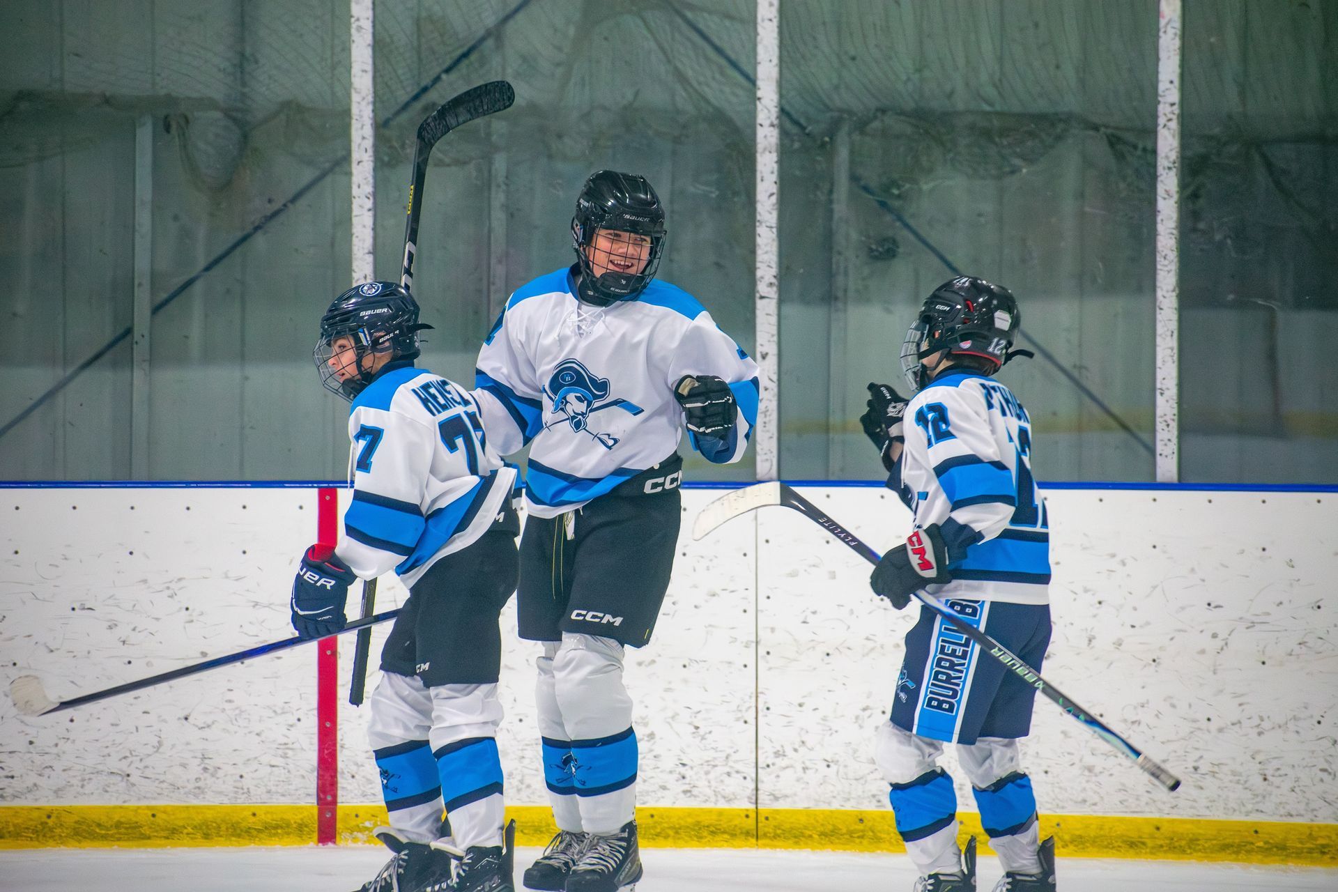 Three ice hockey players in white and blue jerseys celebrate on the ice, smiling and cheering after a play.