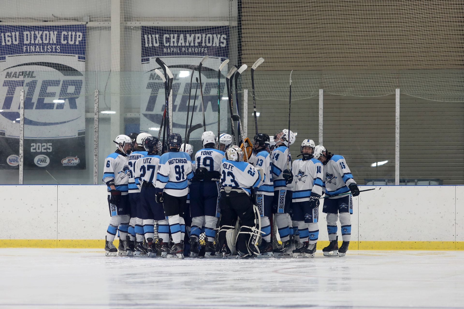 A hockey team in blue and white jerseys huddles together on the ice inside an arena, sticks raised toward the center.