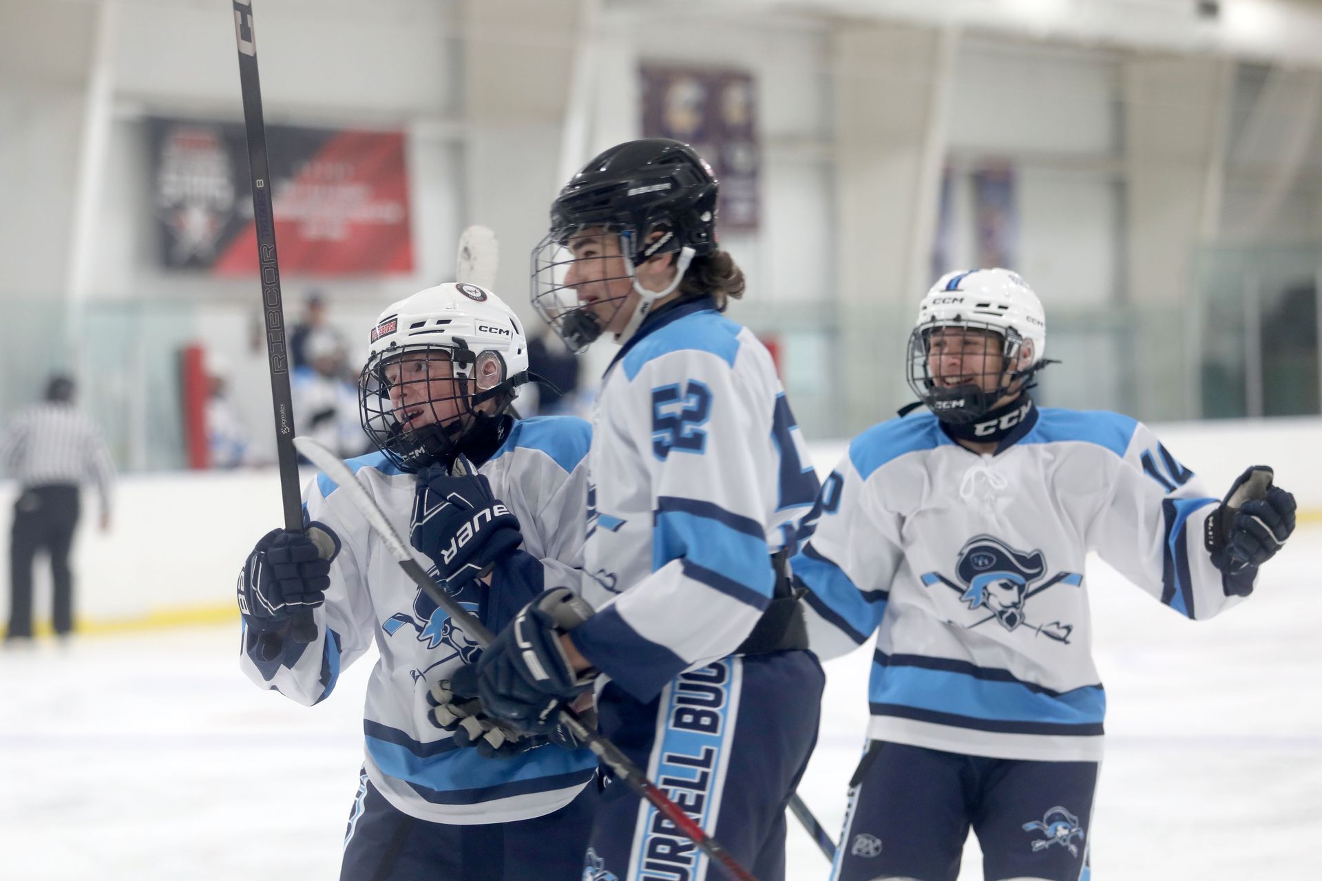 Three ice hockey players in white and blue uniforms celebrate on the ice, smiling and cheering after a goal.