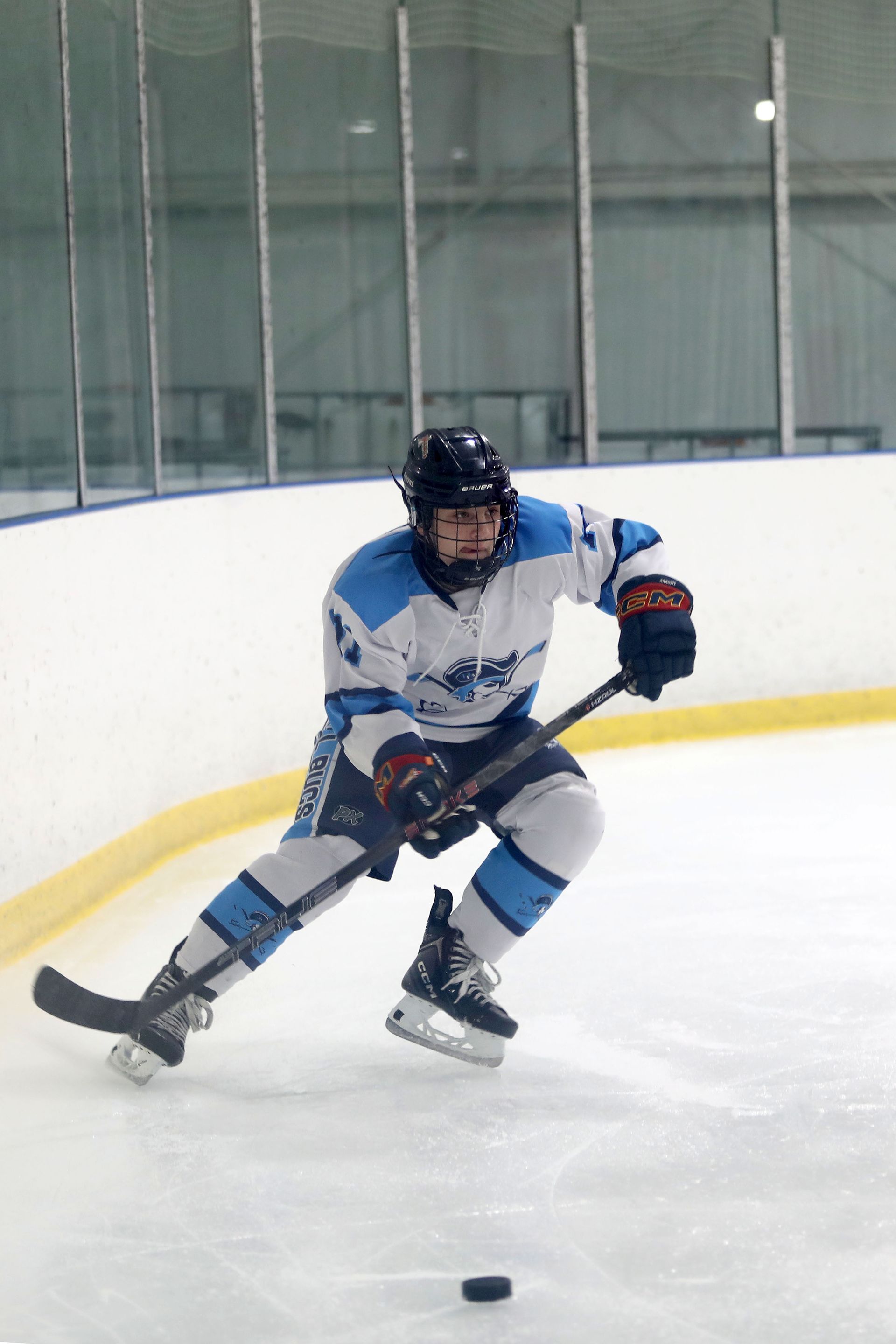 A hockey player in a white and blue jersey skating on an ice rink, maneuvering a puck with a stick.