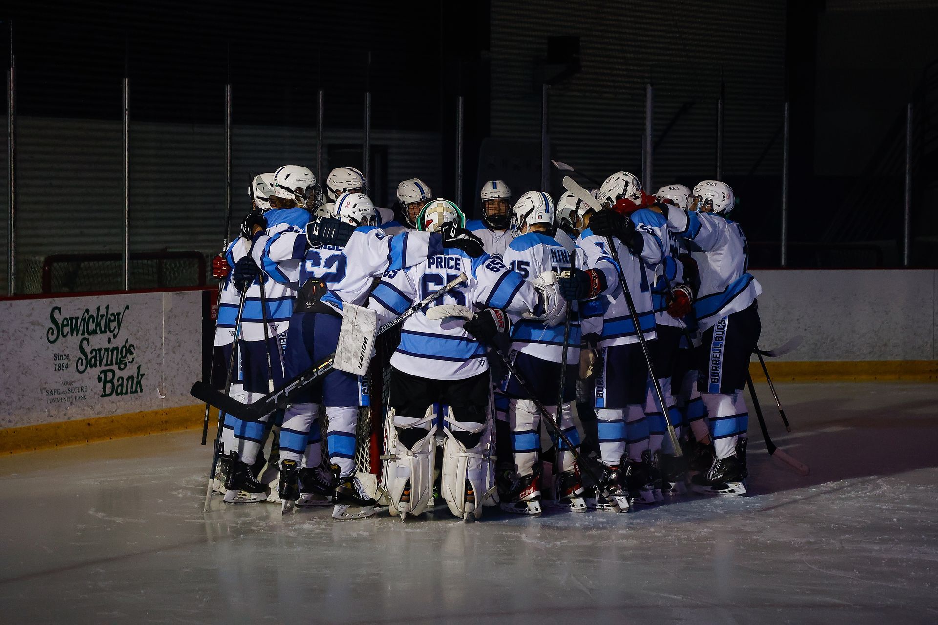 A hockey team in white and blue uniforms stands in a huddle on the ice rink.