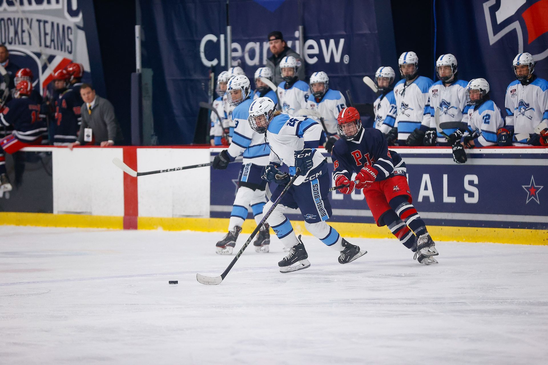 Hockey players in blue and red jerseys compete for control of the puck on an ice rink in front of a team bench.