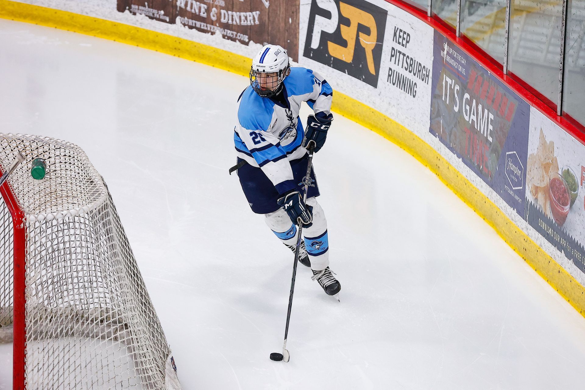 A hockey player in a blue and white jersey skates near the boards, controlling the puck with their stick.