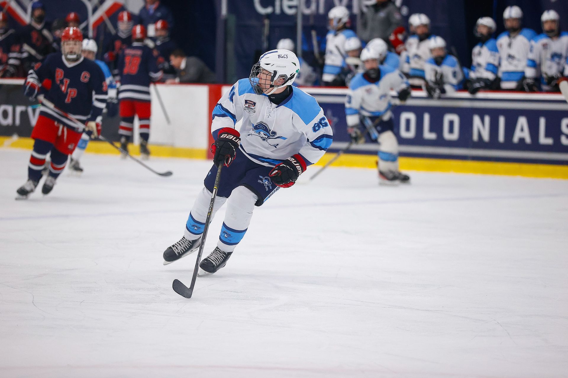 A hockey player in a white and blue uniform skates across the ice, stick in hand, with teammates and opponents nearby.