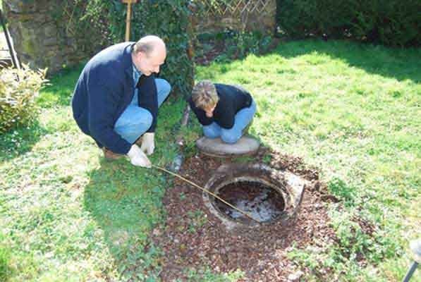 Two people kneel on grass beside an open manhole in a sunny garden.