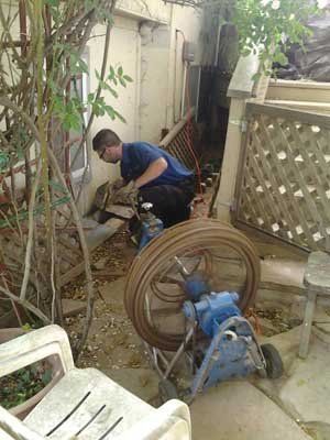 Worker crouching in a narrow outdoor alley, repairing pipes near coiled hoses and a blue pump.