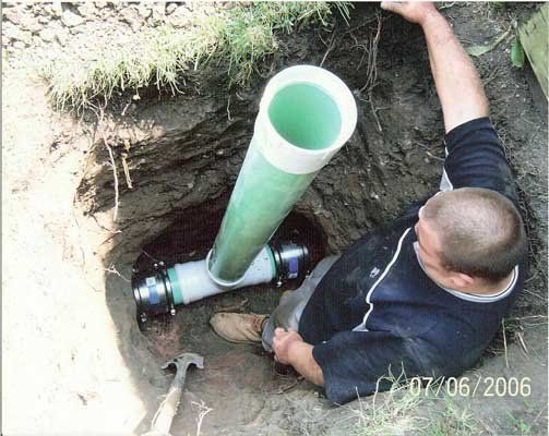 Worker crouches in a dirt trench beside a green pipe connected to a black fitting