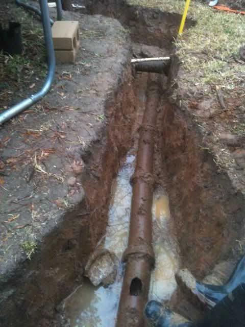 Open trench with exposed underground pipe and muddy soil beside a building foundation