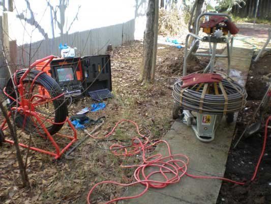 Cluttered outdoor work area with red cables, tools, and a generator under trees and fencing.