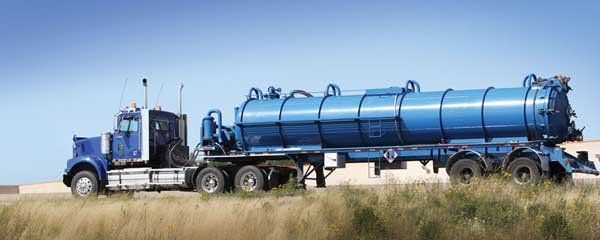 Blue tanker trailer truck driving on a dirt road beside tall grass under a clear sky