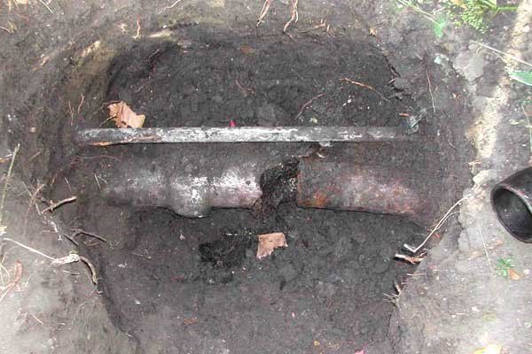 Rusty pipe or valve in a dirt hole outdoors, with a metal bar across the top.