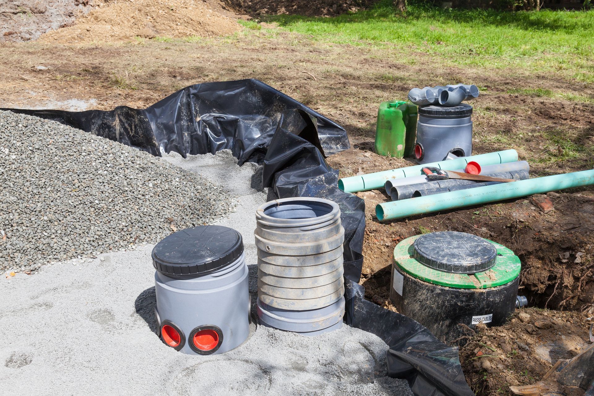 Utility pipes and fittings beside a gravel trench, with a black tarp and a green underground access lid.