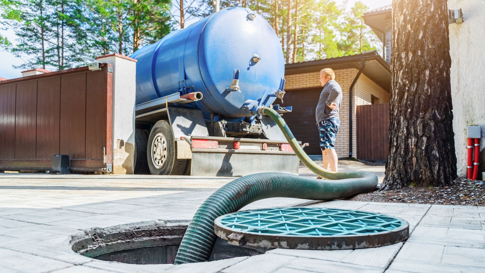 Sewer cleaning truck hoses into open manhole on a sunny street, worker standing nearby Sewer cleaning truck hoses into open manhole on a sunny street, worker standing nearby
