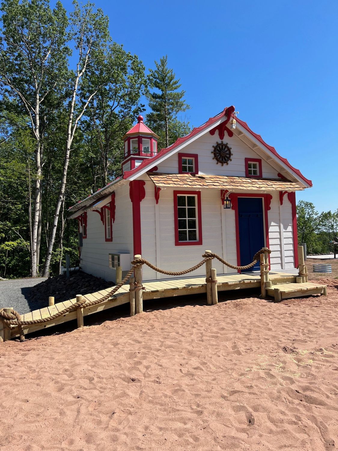 Small white building with red trim, a blue door, and lighthouse-style top, on a wooden platform near sand and trees.