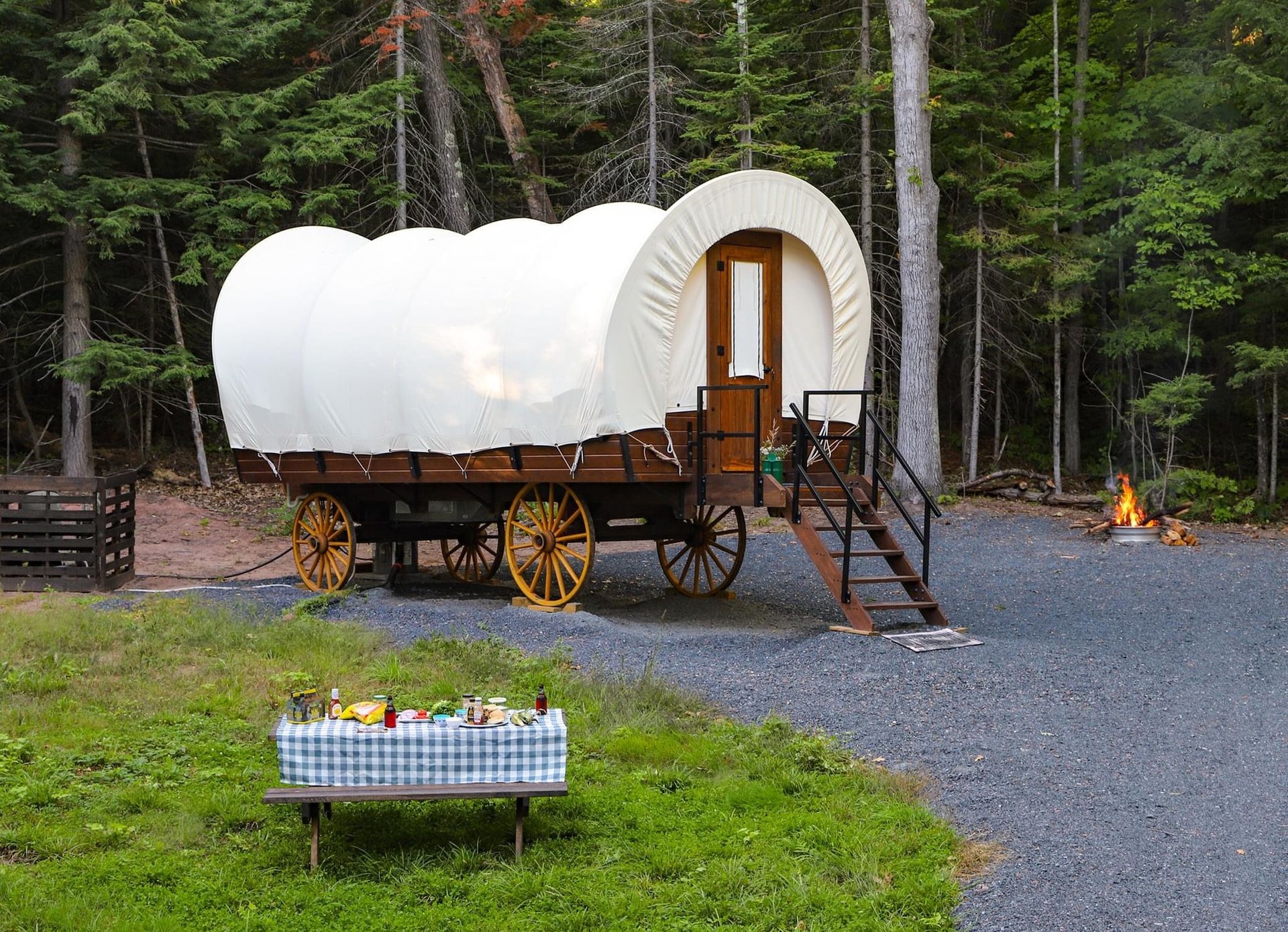 Covered wagon campsite, steps, small picnic table with food/drinks, fire pit, forest background.