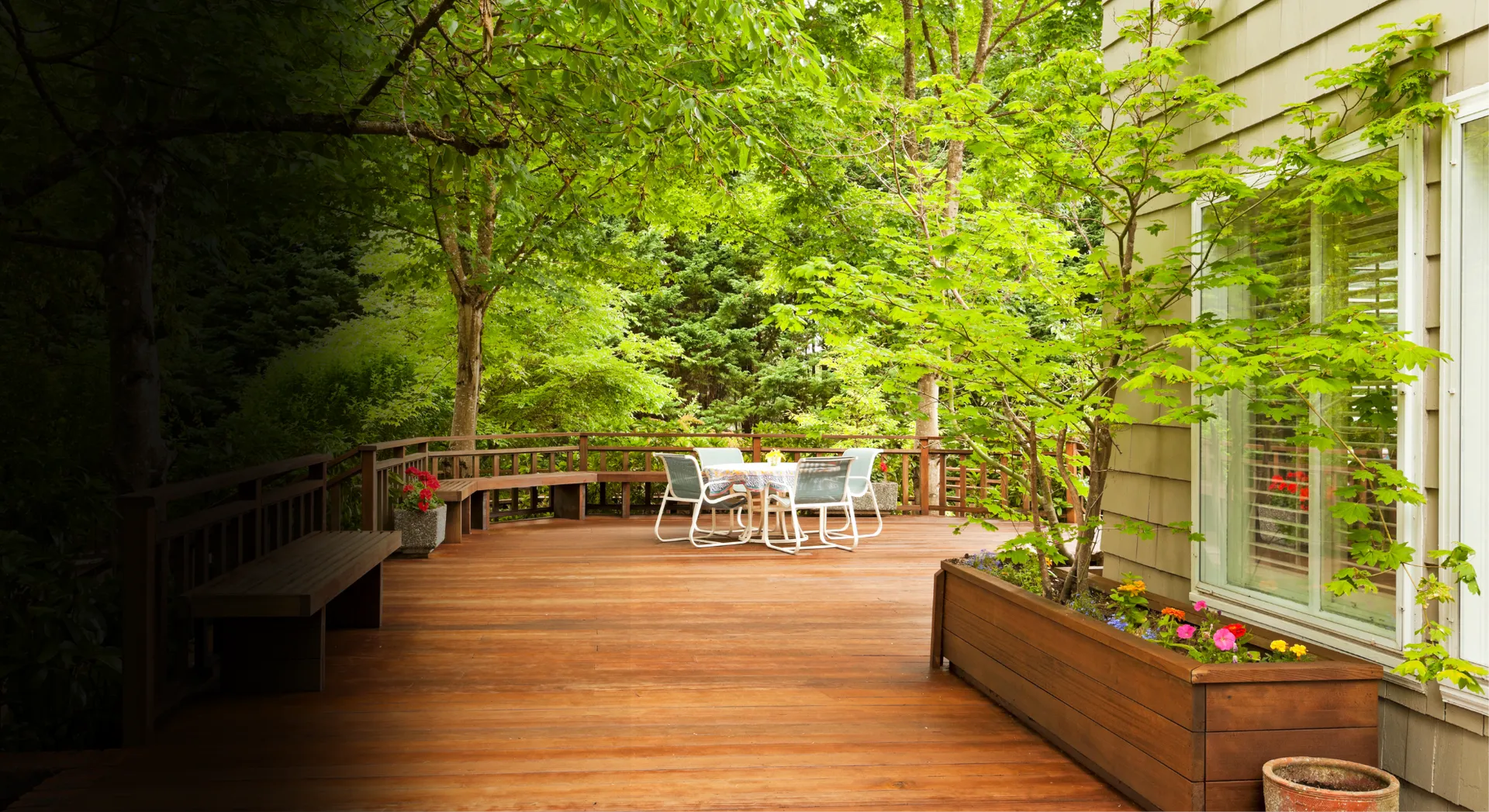 Wooden deck patio with white outdoor table and chairs, surrounded by lush green trees and planters.