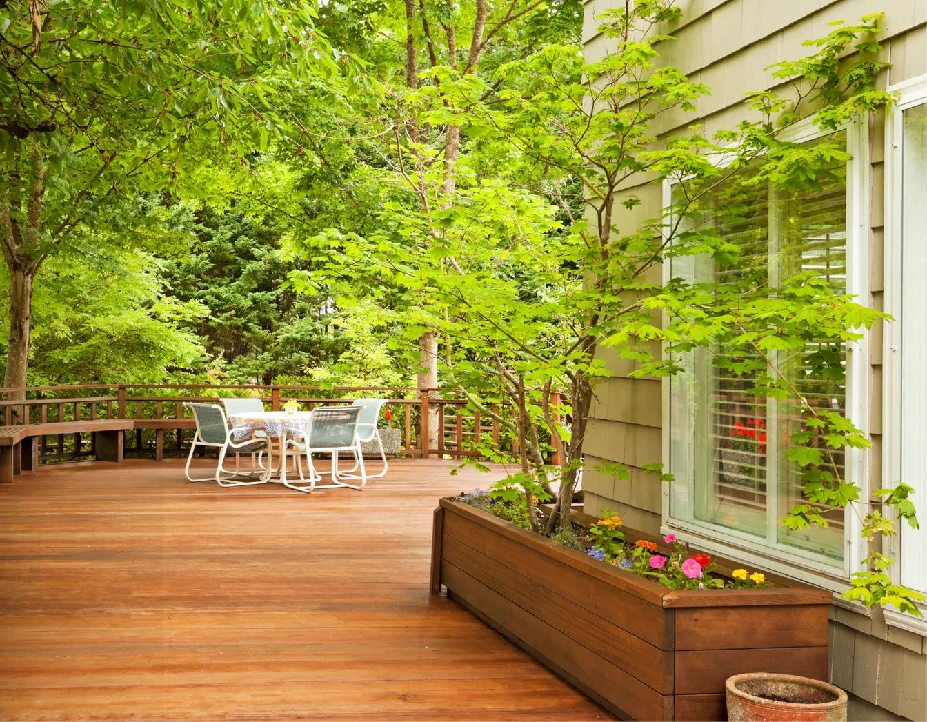 Wooden deck with white patio table and chairs, surrounded by lush green trees and a planter beside house windows.