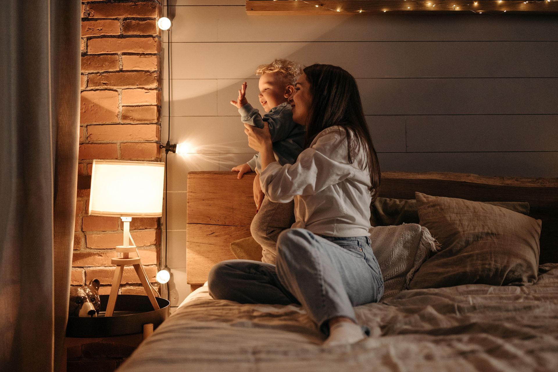 Couple cuddling on a bed in a cozy, warmly lit bedroom with a bedside lamp and brick wall