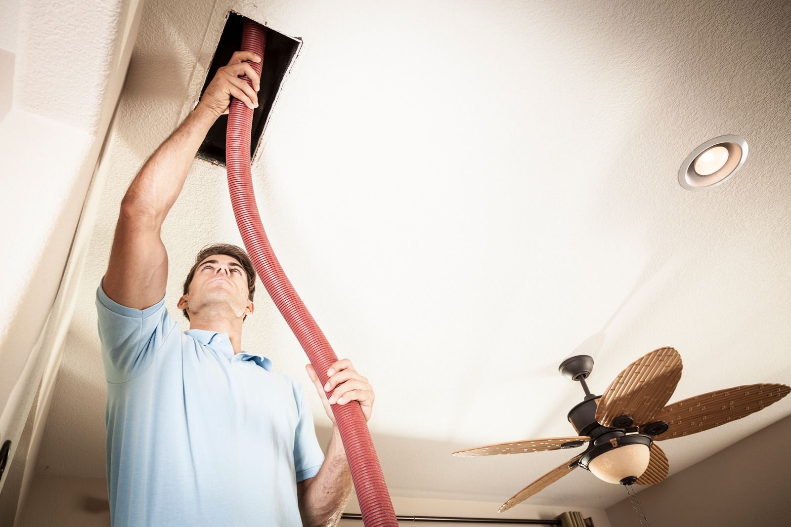 Man in a blue shirt vacuuming dust from a ceiling vent with a red hose under a ceiling fan