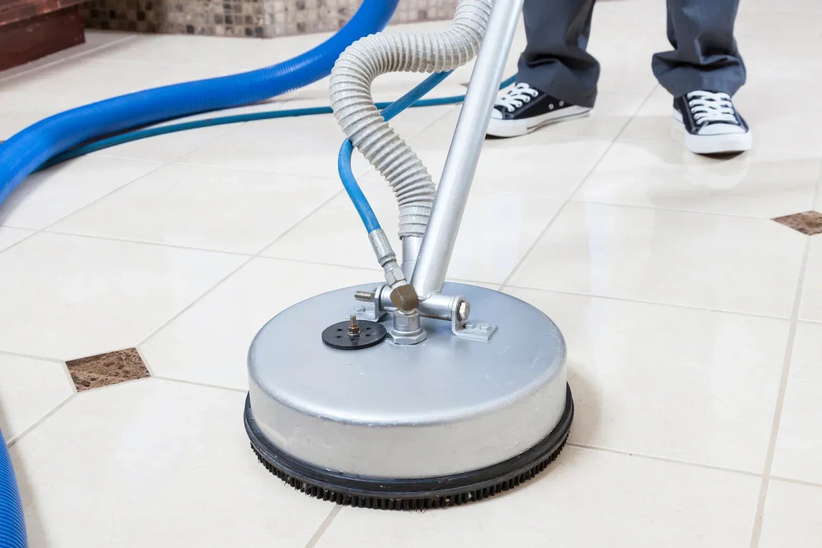 Floor cleaning machine scrubbing white tile floor, with hoses attached and a person standing nearby